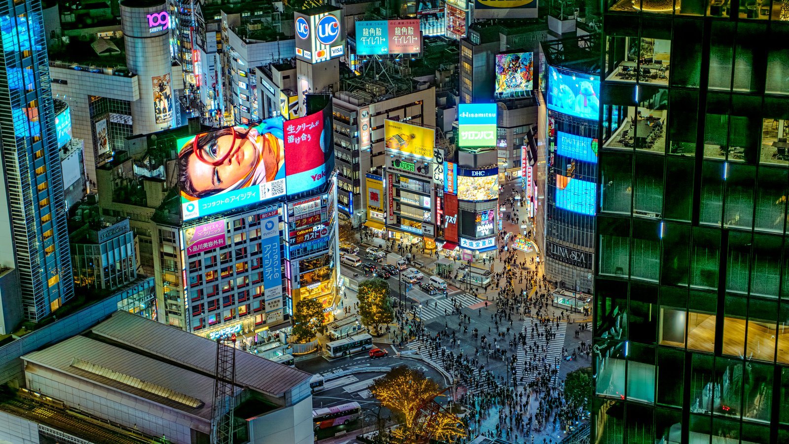Shibuya Crossing in Tokyo seen from above at night, crowds moving through a canyon of lit screens